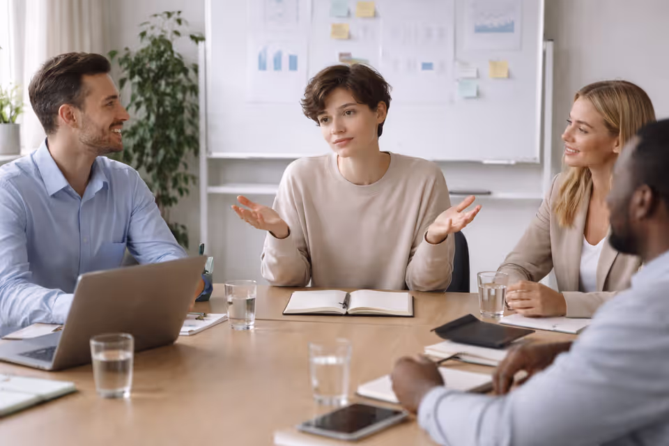 A person apologizing unnecessarily during a workplace meeting while colleagues look unconcerned